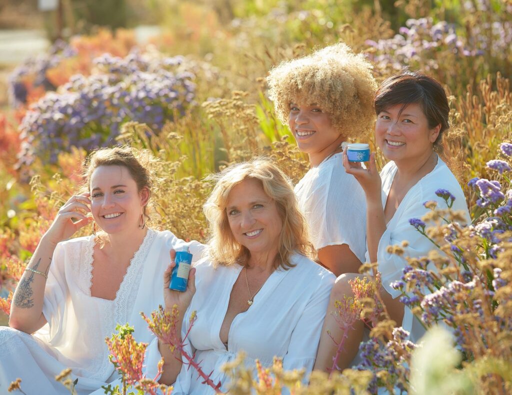 Models Ariana Wohlstattar, Mary Riley, Kéme Chambers and Lisa Amorão hold glass bottles of clean beauty brand Green Bee Botanicals' Fresh Face Toner and Quench Moisturizer. The models are wearing white dresses, sitting in a field of wildflowers at Sonoma Hills Farm in Petaluma, California. 