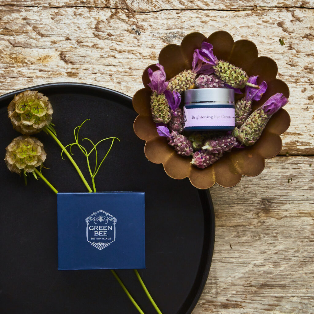 Green Bee Botanicals Brightening Eye Cream box and jar nestled on vintage trays with cannabis and other flowers against rustic wood